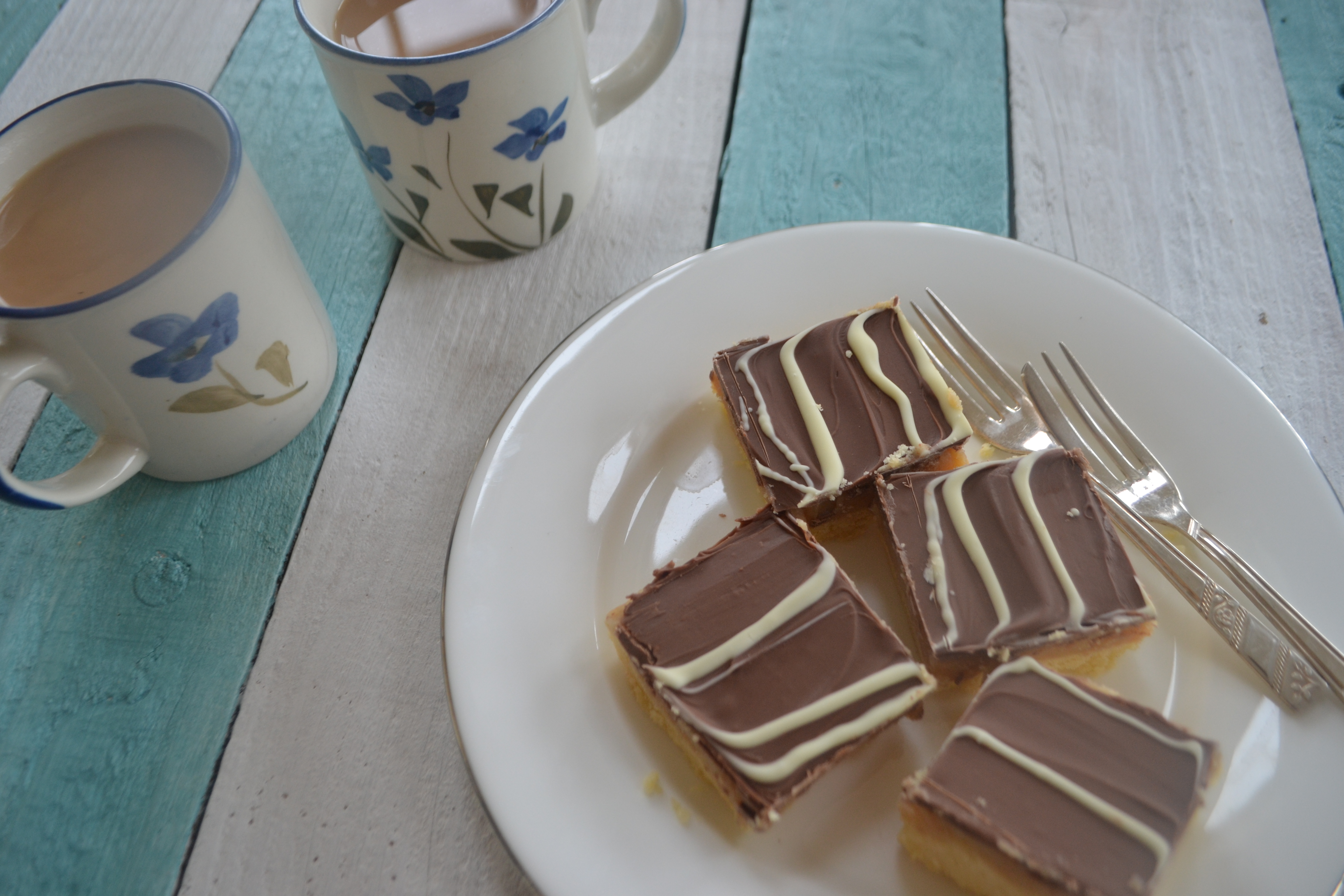 A plate with 4 pieces of Millionaire's shortbread, beside 2 teacups